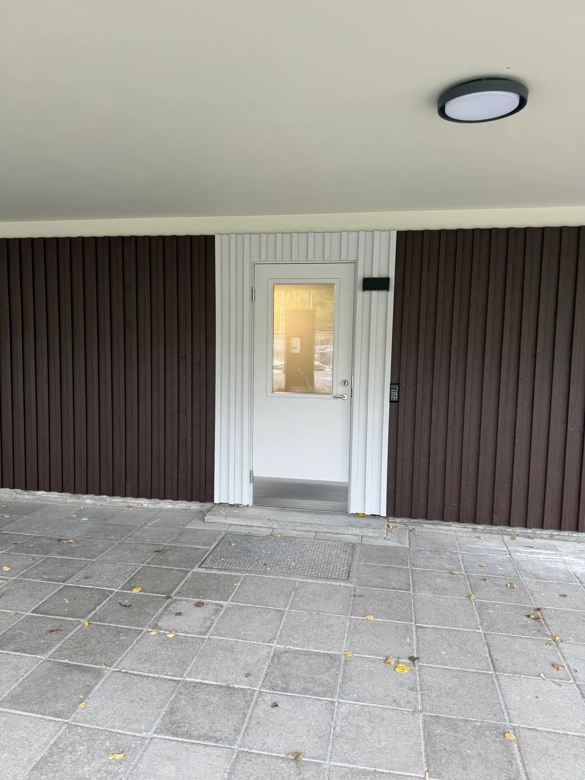 Entré med mörk träpanel och ljus dörr, plattlagd markyta framför